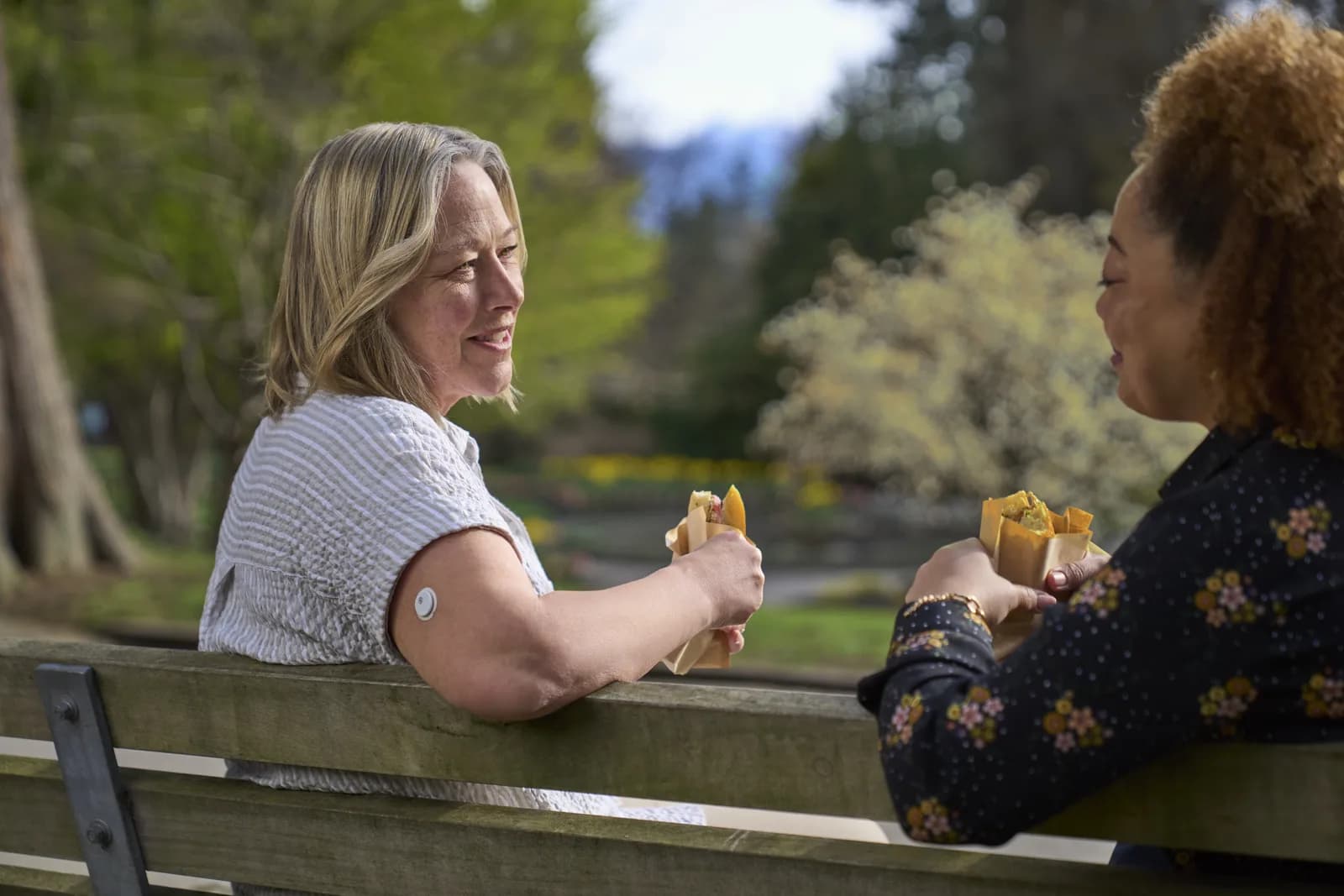Two friends talking on a park bench, one wearing a glucose sensor on her upper arm